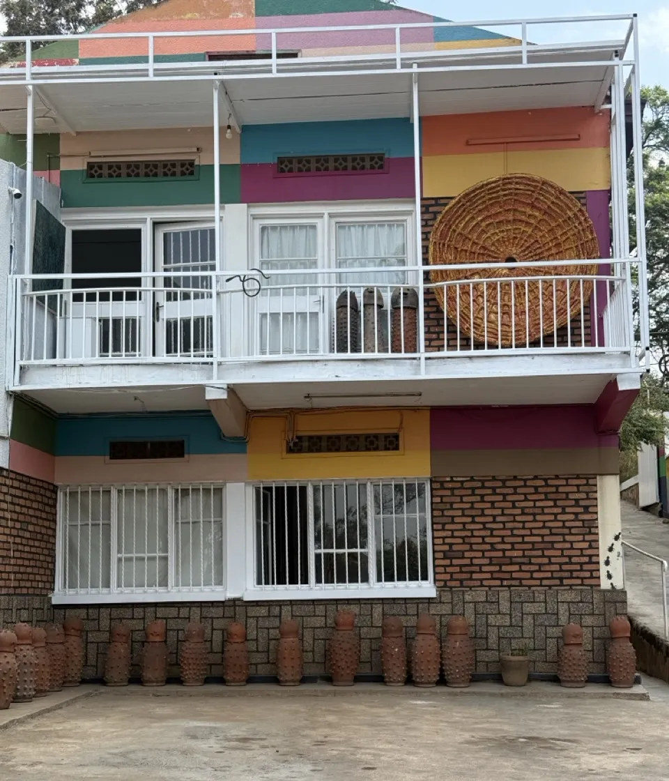 Two-storey building painted in colour-block stripes, with a large woven disc on the balcony wall and a row of studded terracotta pots along the front