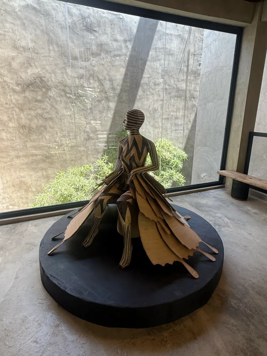 Sculpture of a seated figure with black and natural zigzag patterns on a round black plinth, in a gallery by a tall window